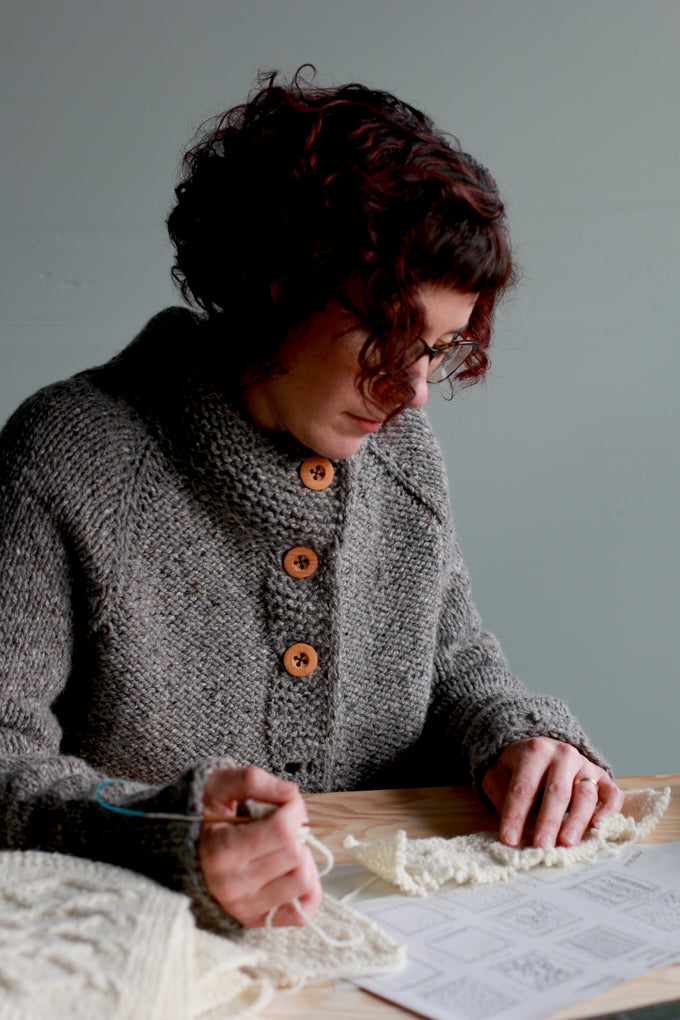 Carol Feller wearing the 'Sorn' cardigan, sitting at a desk looking at a swatch of Irish Galway Sheep yarn.